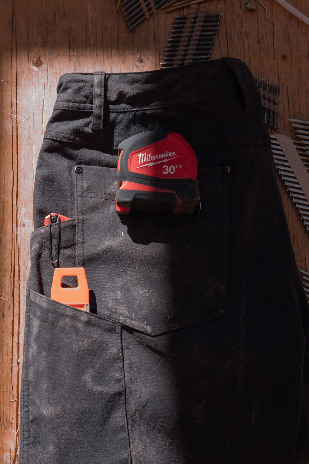 The 686 Men's Unwork® Pant - Relaxed Loose Fit lies on a wooden floor with a Milwaukee tape measure and orange utility knife in the back pocket. Nails and dust are scattered around these durable 686 work pants.