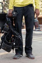 Wearing 686 Men's Unwork® Pant - Relaxed Loose Fit, a person in classic workwear stands at a worksite, holding a tool belt and drink can. Only their lower body is visible.
