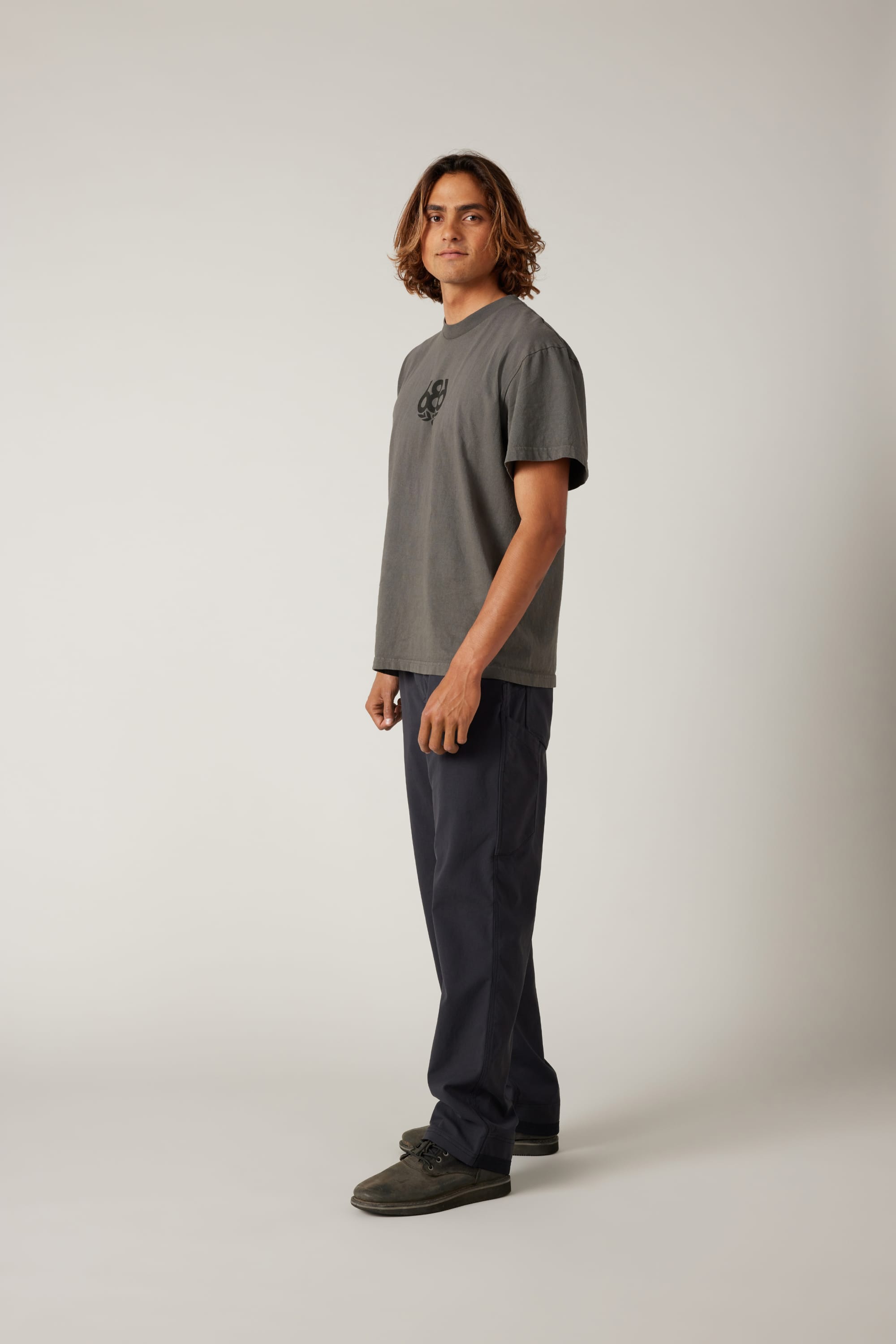 A young man with shoulder-length brown hair stands in a studio, wearing the 686 Men's Unwork® Pant - Relaxed Loose Fit and a gray T-shirt, looking at the camera with a neutral expression against a plain light background.