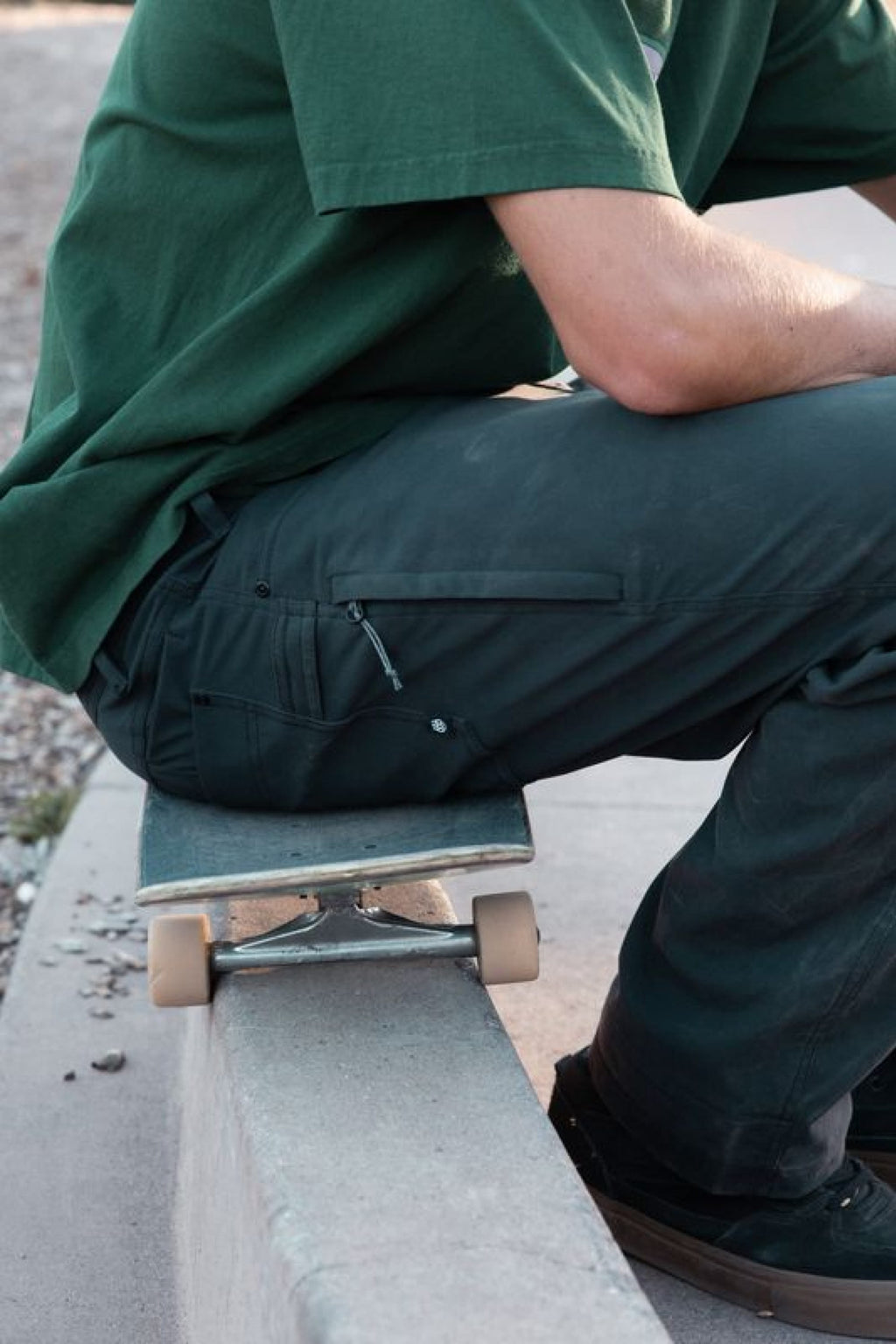 Wearing the 686 Men's Unwork® Pant - Relaxed Loose Fit, a person sits on a skateboard atop a concrete ledge outdoors. Their face is not visible in the image.