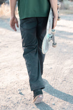 A person in 686 Men's Unwork® Pant - Relaxed Loose Fit walks away on a sunlit path, holding a skateboard by their side. Their green shirt stands out, while their face remains unseen in the image.