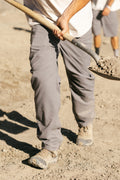 Wearing 686 Men's Unwork® Pant - Relaxed Loose Fit, someone in classic workwear and tan boots uses a shovel on sandy ground, with another person blurred in the background.