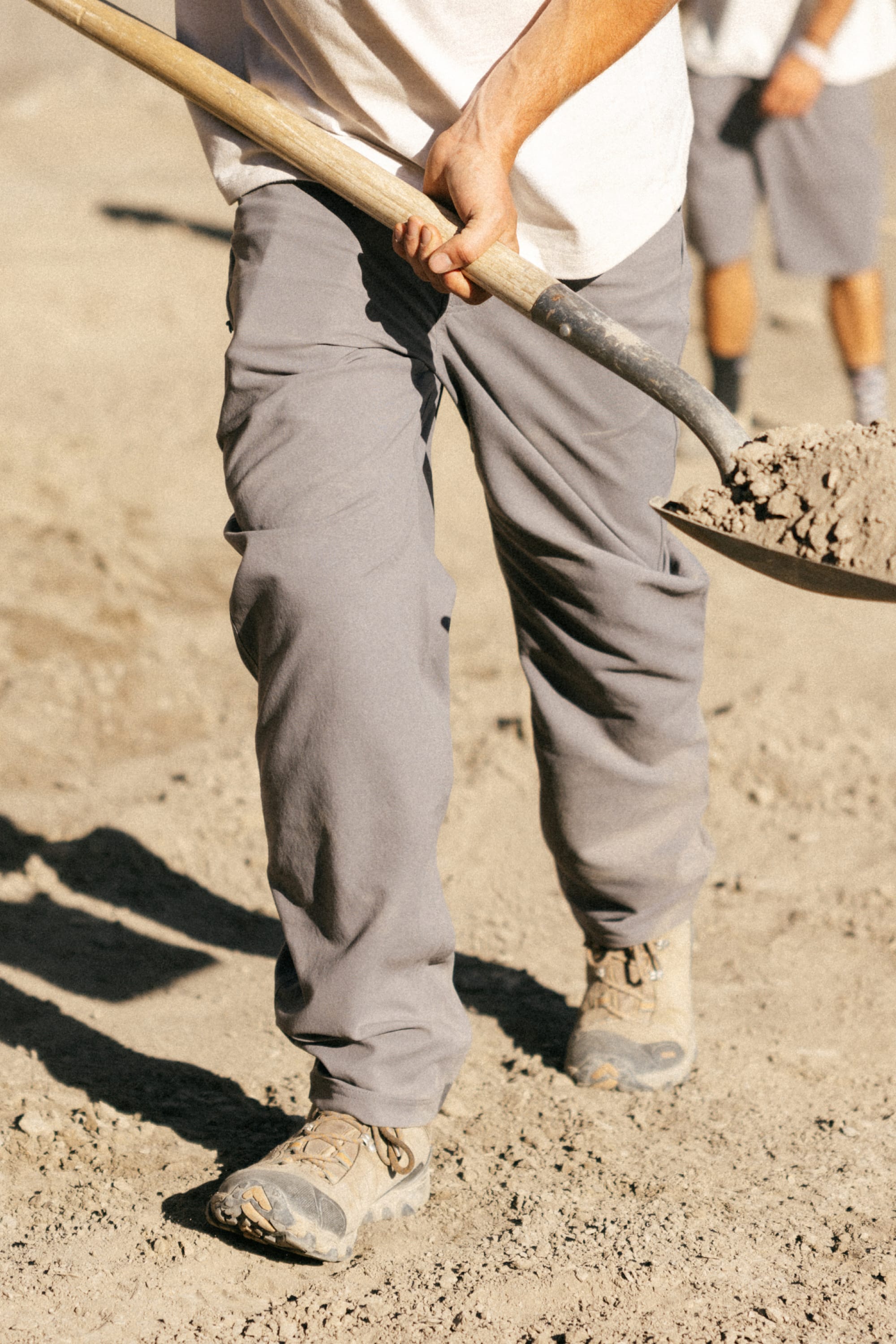 Wearing 686 Men's Unwork® Pant - Relaxed Loose Fit, someone in classic workwear and tan boots uses a shovel on sandy ground, with another person blurred in the background.