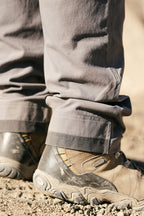 A close-up of someone outdoors in 686 Men's Unwork® Pant - Relaxed Loose Fit by 686, the gray pants resting slightly wrinkled over light brown hiking boots on a sandy, rocky surface.
