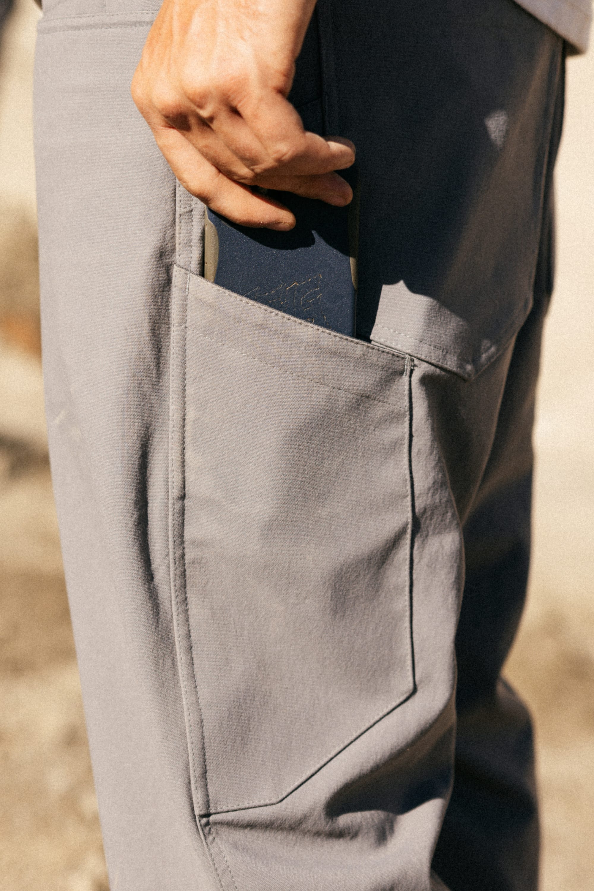 A close-up of someone slipping a dark notebook into the side pocket of 686 Men's Unwork® Pant - Relaxed Loose Fit. The sunlit background is blurred, highlighting the pant's practical design and durability.