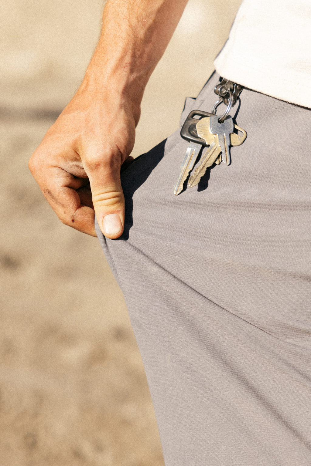 A hand pulls the pocket of the 686 Men's Unwork® Pant - Relaxed Loose Fit, with a keychain clipped to the durable waistband. The classic workwear-inspired gray pants stand out against a blurred background.