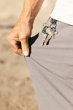 A hand pulls the pocket of the 686 Men's Unwork® Pant - Relaxed Loose Fit, with a keychain clipped to the durable waistband. The classic workwear-inspired gray pants stand out against a blurred background.