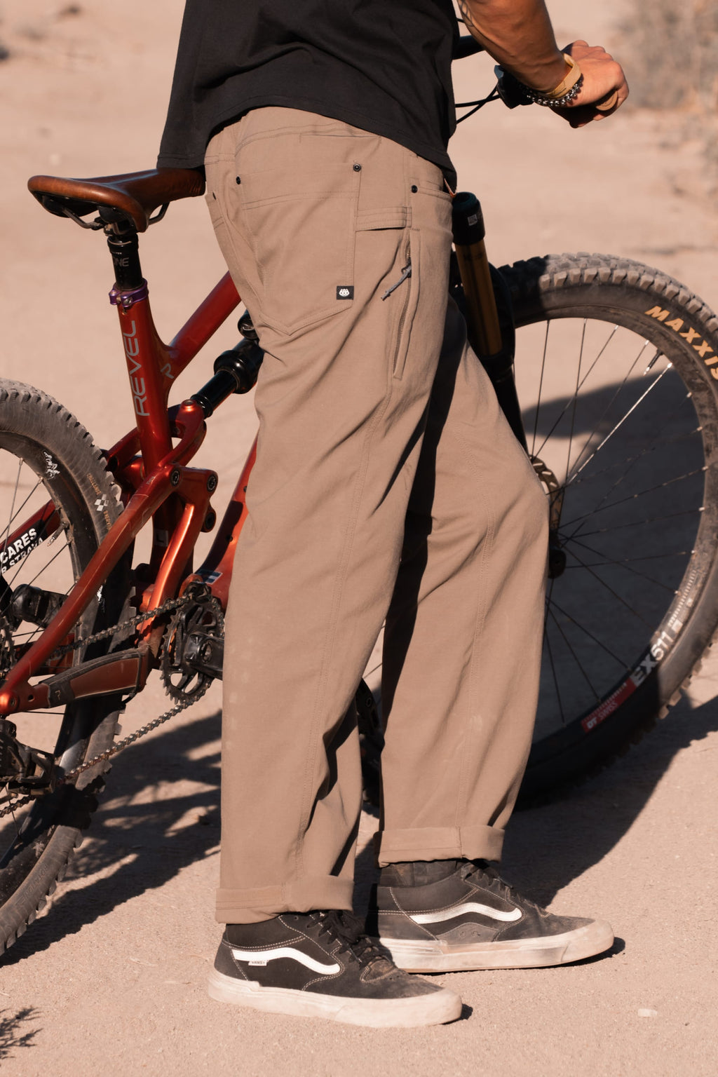 A person wearing 686 Men's Unwork® Pant - Relaxed Loose Fit stands next to a red mountain bike on a dirt path, holding the handlebars. Only their lower body is visible.