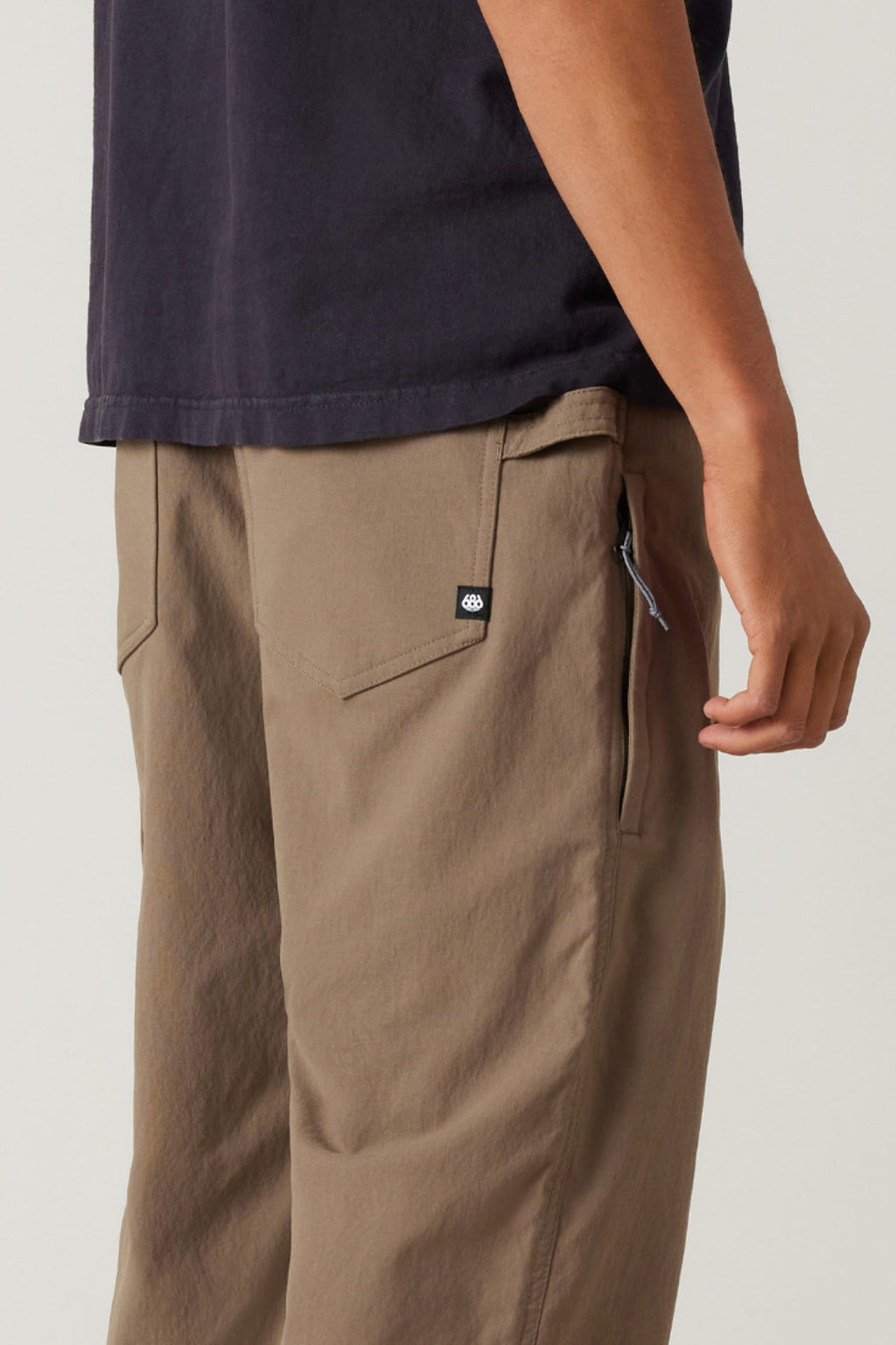 A person shown from the waist down and side, wearing a dark t-shirt with 686 Men's Unwork® Pant - Relaxed Loose Fit by 686: light brown, durable pants featuring a zippered pocket with flap.