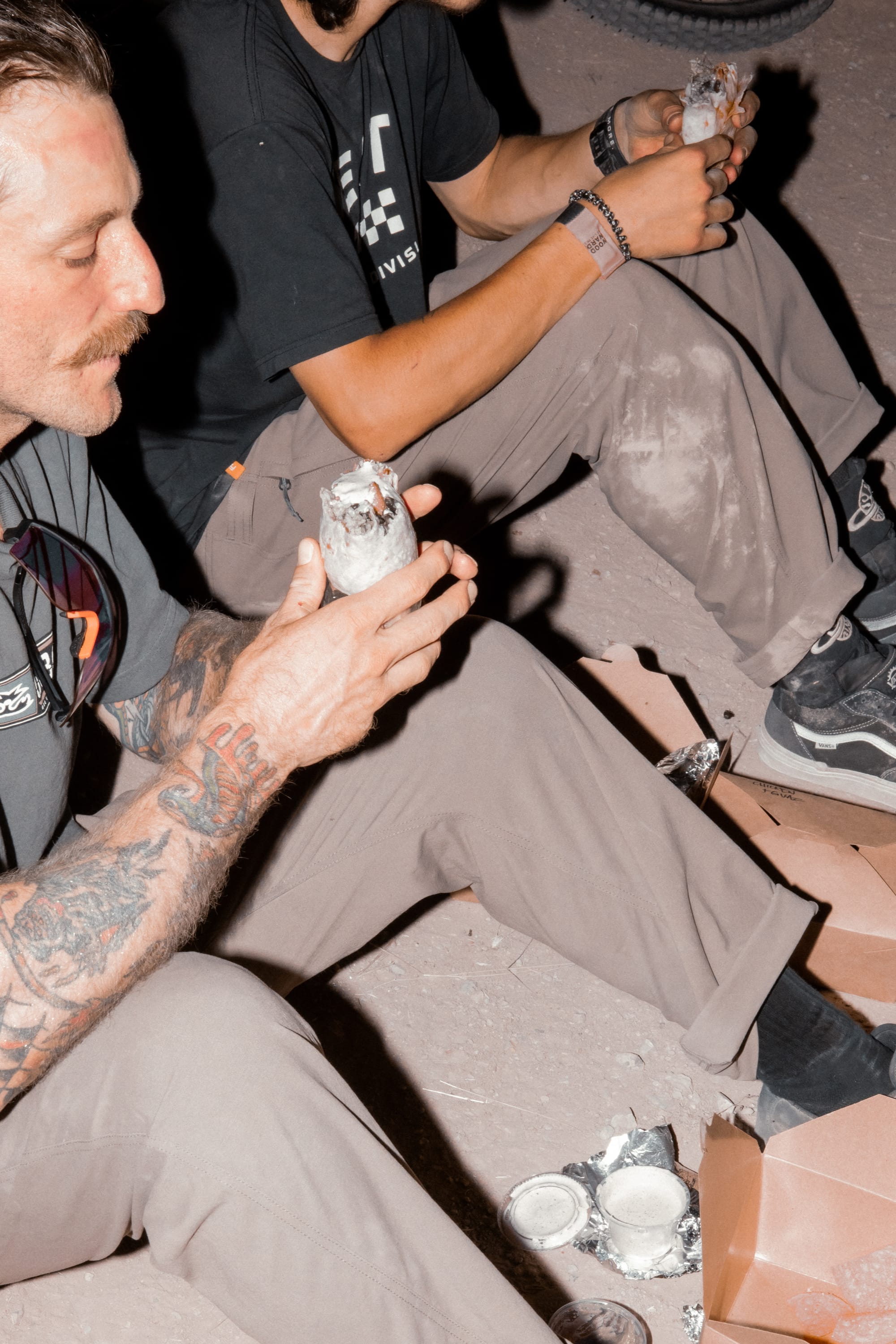Two people in casual outdoor clothing, including 686 Men's Unwork® Pant - Relaxed Loose Fit by 686, sit on the ground eating foil-wrapped burritos among takeout containers; one has tattooed arms and a mustache.
