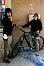 Two men in 686® Men's Unwork® Pant - Relaxed Loose Fit stand by a bicycle, showcasing 686's blend of comfort and durability for outdoor adventures.