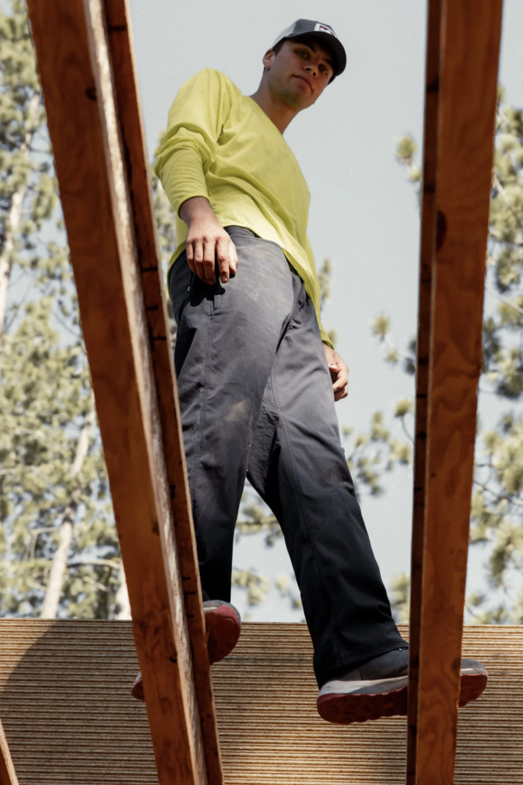 Wearing a yellow shirt, a classic workwear cap, and 686 Men's Unwork® Pant - Slim Fit, a person walks on wooden beams at a construction site with trees and sky in the background. The upward photo angle emphasizes the scene.