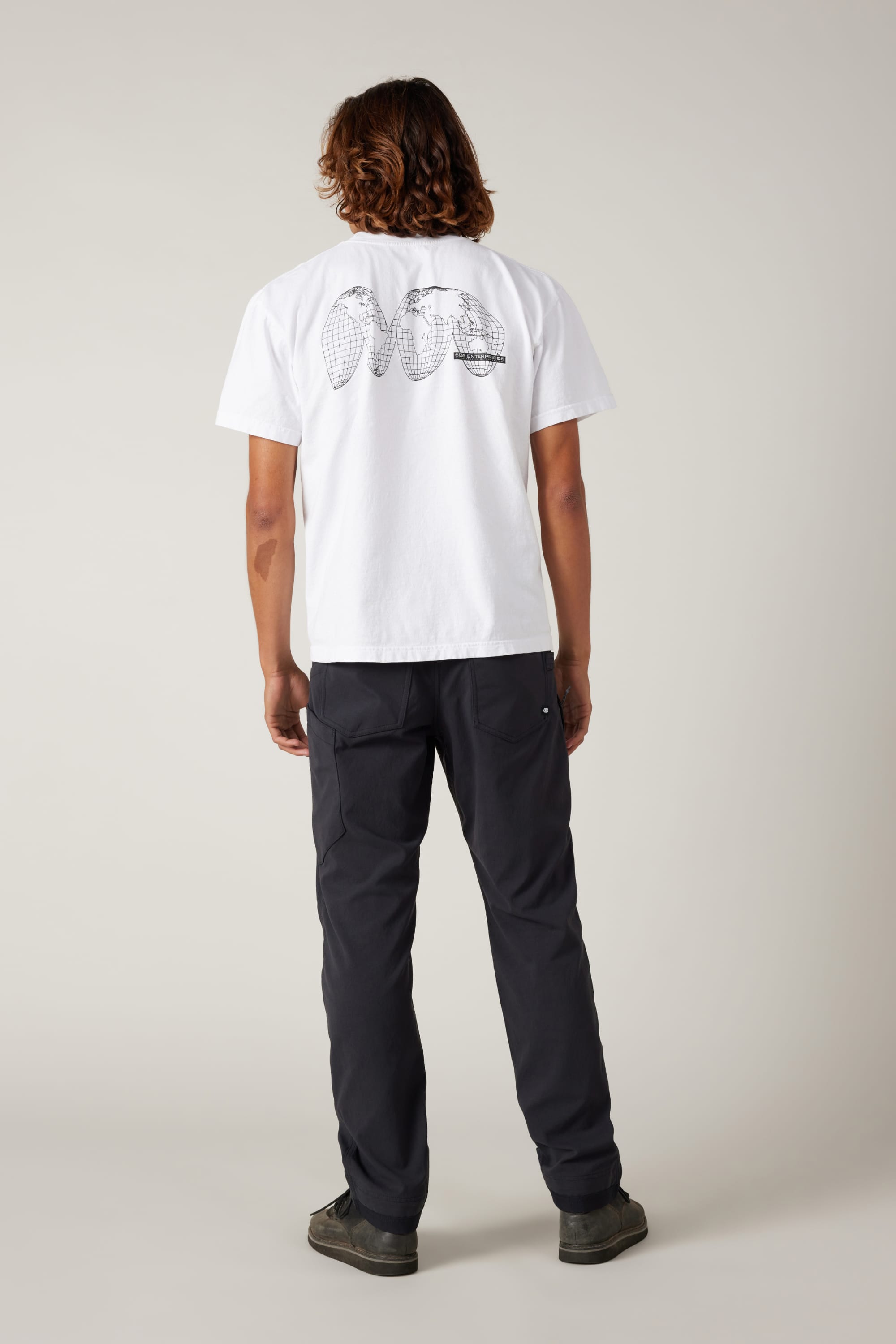 A person with shoulder-length hair stands facing away in a white tee with a black back graphic, 686 Men's Unwork® Pant - Slim Fit by 686 in dark color, and dark shoes, set against a plain light background.