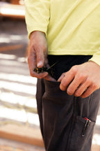 A person in a yellow long-sleeve shirt holds several nails while reaching into the pocket of their 686 Men's Unwork® Pant - Slim Fit, with a pencil visible in the pocket.