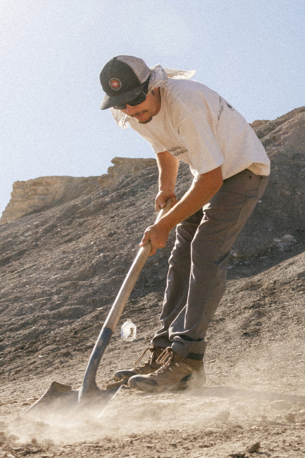 Wearing a cap, sunglasses, and 686 Men's Unwork® Pant - Slim Fit, someone uses a shovel to dig in dry ground under the sun, with rocky hills behind them.