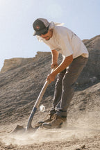 Wearing a cap, sunglasses, and 686 Men's Unwork® Pant - Slim Fit, someone uses a shovel to dig in dry ground under the sun, with rocky hills behind them.