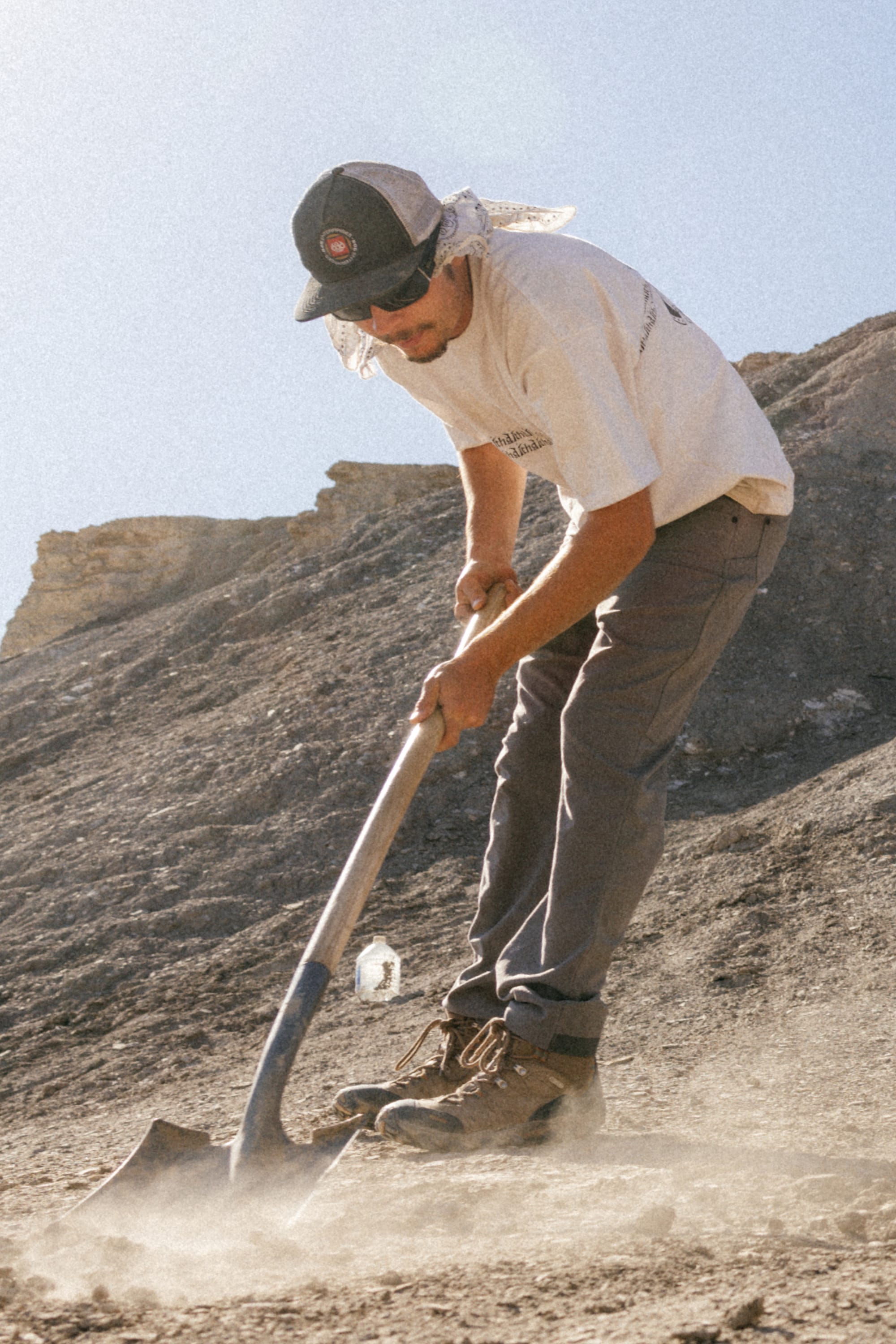 Wearing a cap, sunglasses, and 686 Men's Unwork® Pant - Slim Fit, someone uses a shovel to dig in dry ground under the sun, with rocky hills behind them.