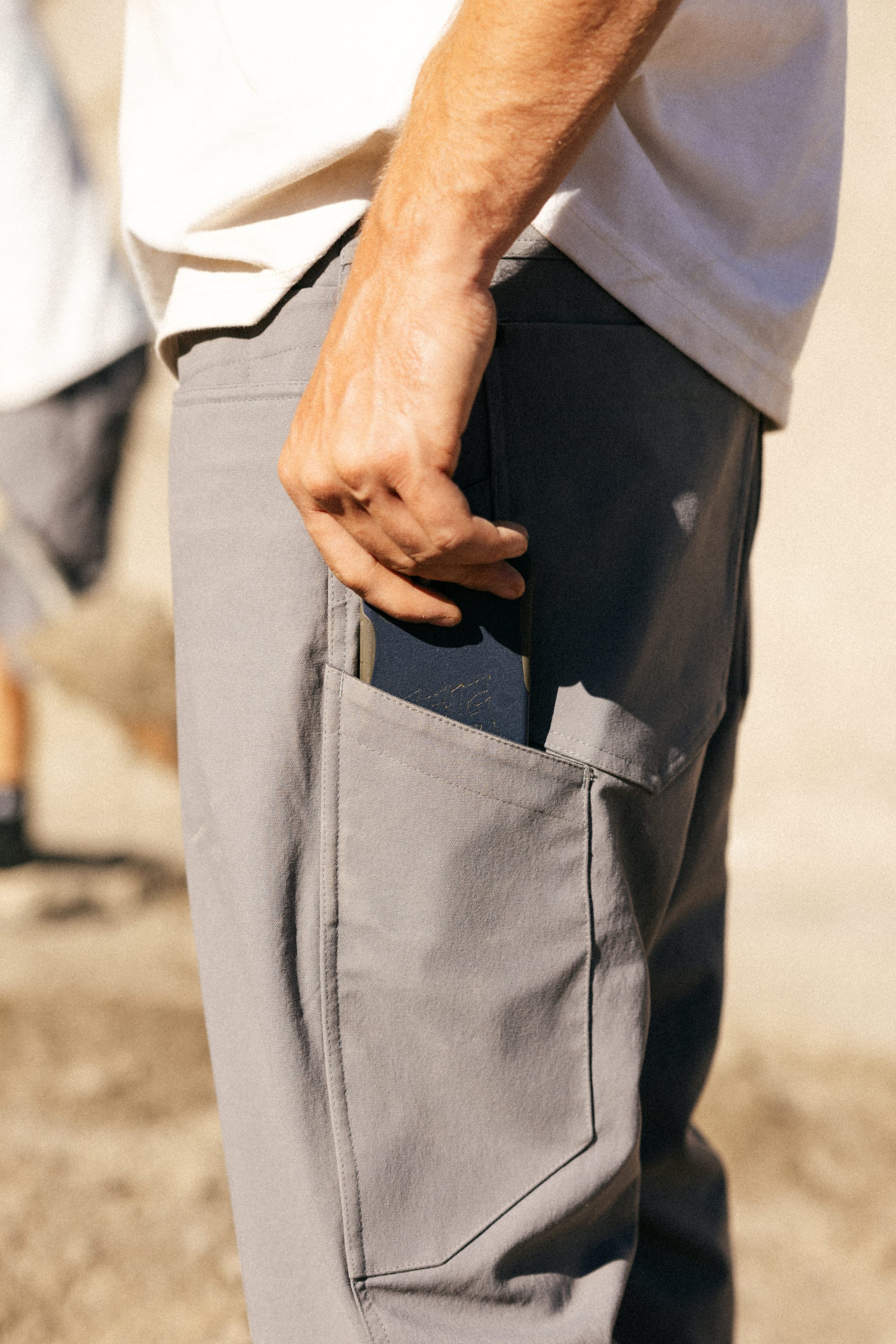 A person wearing 686 Men's Unwork® Pant - Slim Fit in gray and a white shirt slides a black smartphone into the side pocket. The outdoor image is taken in bright sunlight.