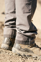 Close-up of a person wearing 686 Men's Unwork® Pant - Slim Fit and rugged hiking shoes on dry, sandy ground. The gray pants by 686 are durable, while the shoes' textured soles and stitching make them ideal for outdoor adventures.