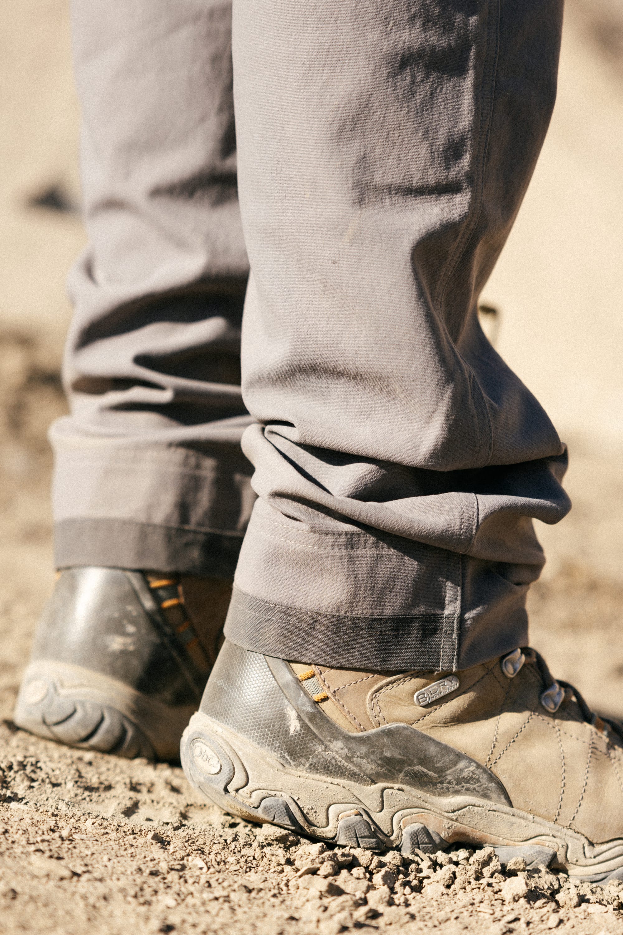 Close-up of a person wearing 686 Men's Unwork® Pant - Slim Fit and rugged hiking shoes on dry, sandy ground. The gray pants by 686 are durable, while the shoes' textured soles and stitching make them ideal for outdoor adventures.