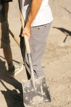 A person wearing 686 Men's Unwork® Pant - Slim Fit in gray and a white shirt holds a shovel on sandy ground, while another person in shorts and sneakers is partially visible beside them.