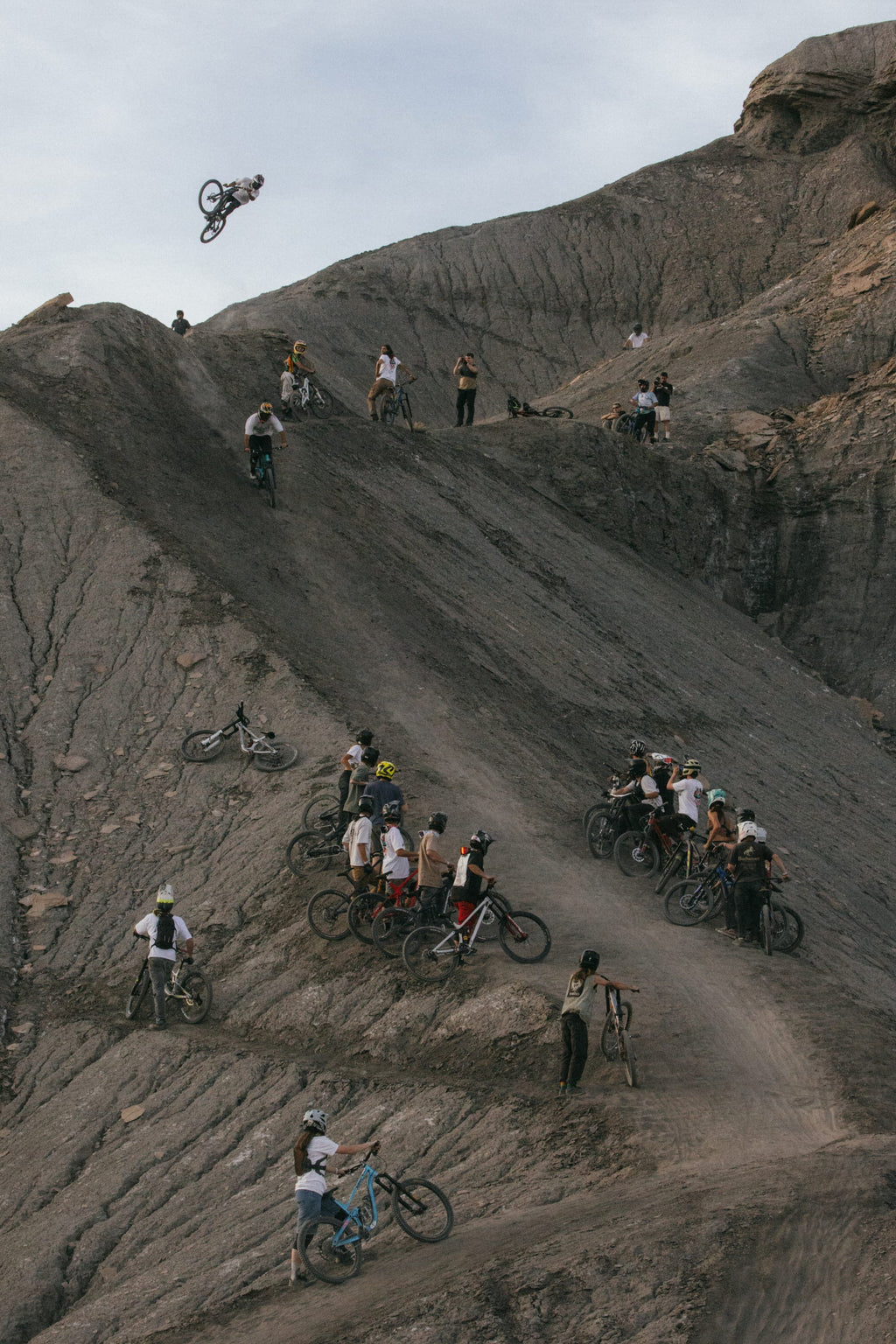 A group of mountain bikers in 686 Men's Unwork® Pant - Slim Fit by 686 watch as a rider executes a high jump from a steep, rocky slope, set amid rugged terrain and dramatic cliffs under a cloudy sky.