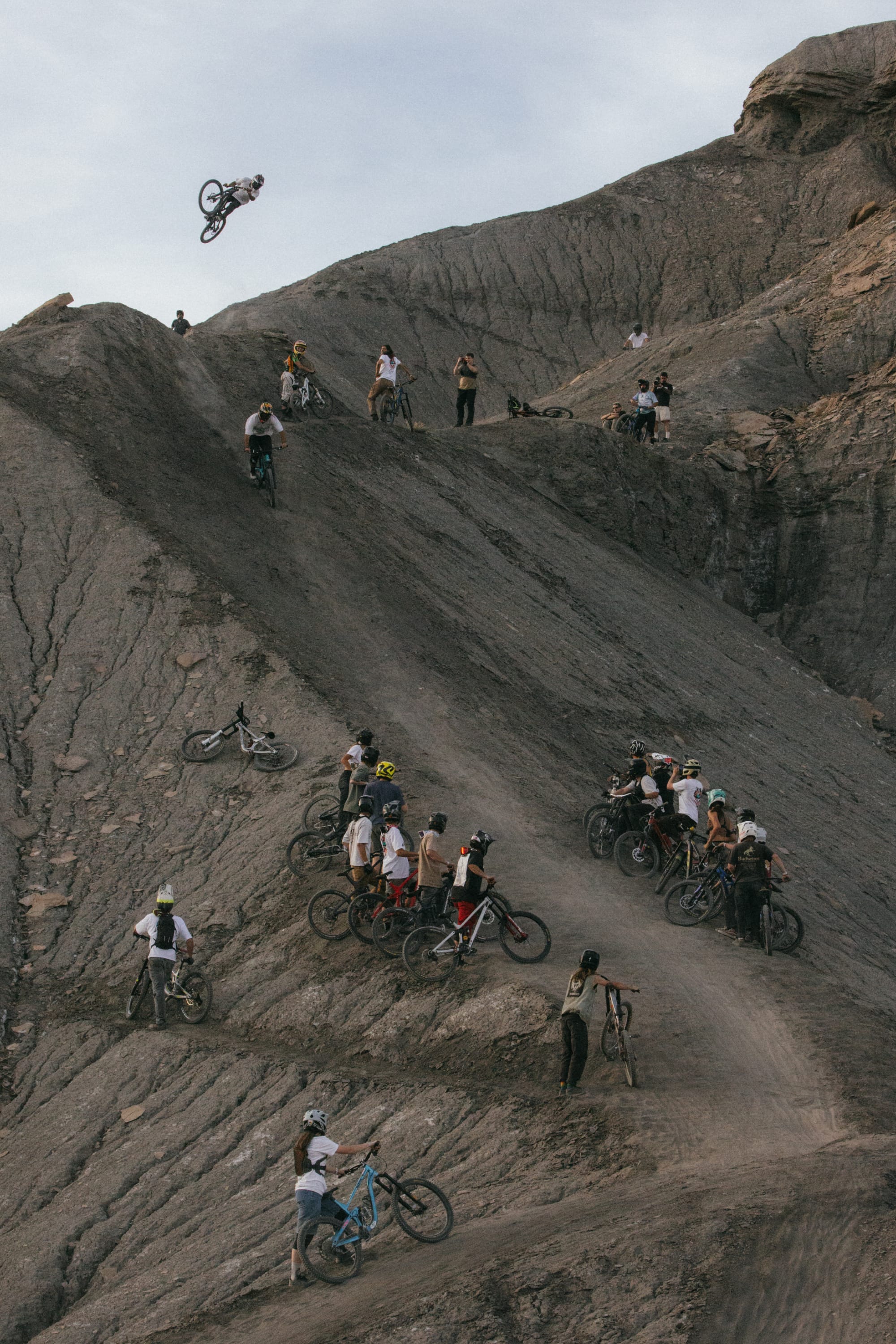 A group of mountain bikers in 686 Men's Unwork® Pant - Slim Fit by 686 watch as a rider executes a high jump from a steep, rocky slope, set amid rugged terrain and dramatic cliffs under a cloudy sky.