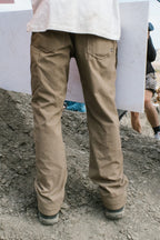 A person wearing 686 Men's Unwork® Pant - Slim Fit stands on rocky ground with a white sign in front; others and outdoor scenery appear in the background.