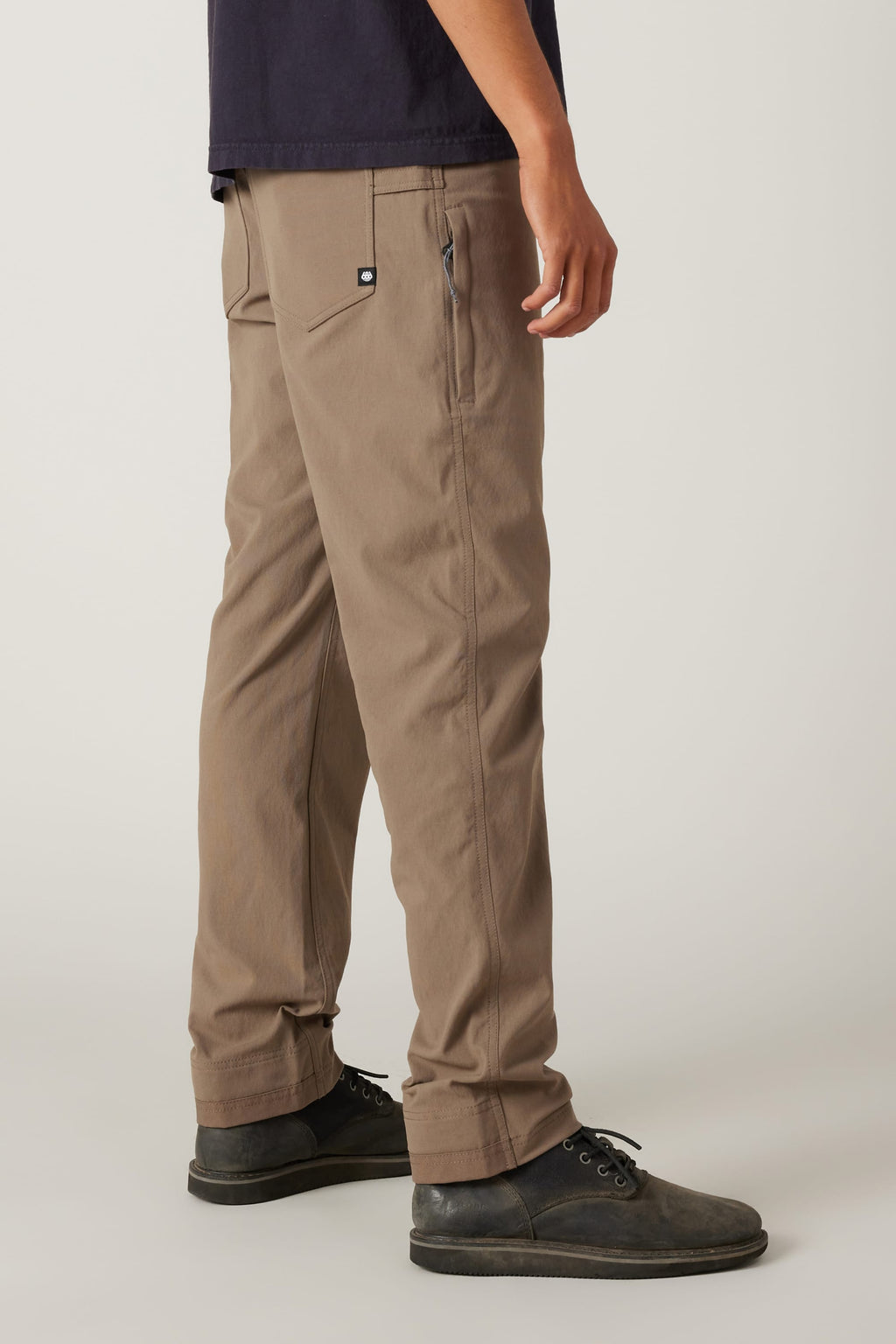 A person stands sideways against a plain background wearing the 686 Men's Unwork® Pant - Slim Fit in brown, black lace-up shoes, and a dark shirt, with their right hand relaxed at their side.