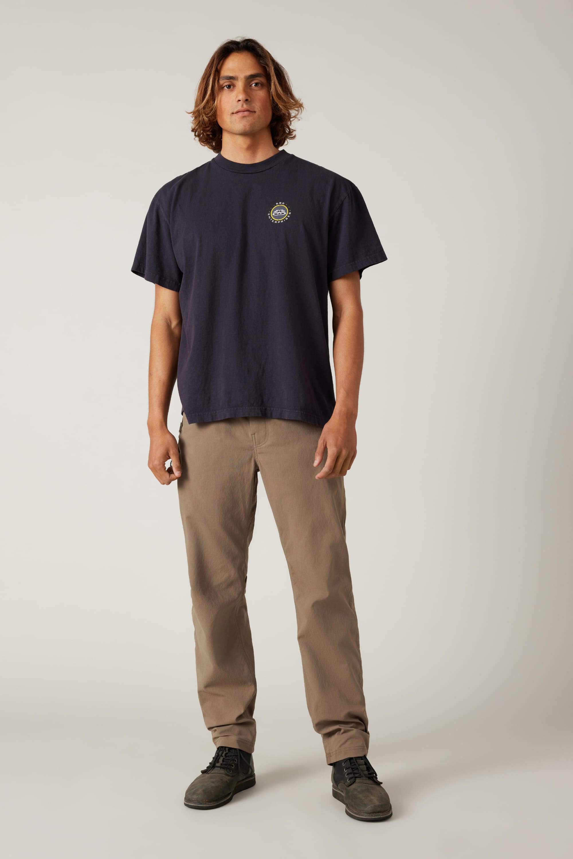 A man with shoulder-length brown hair stands facing forward, wearing a navy blue T-shirt and 686 Men's Unwork® Pant - Slim Fit in classic beige by 686, plus black shoes, set against a plain light background.