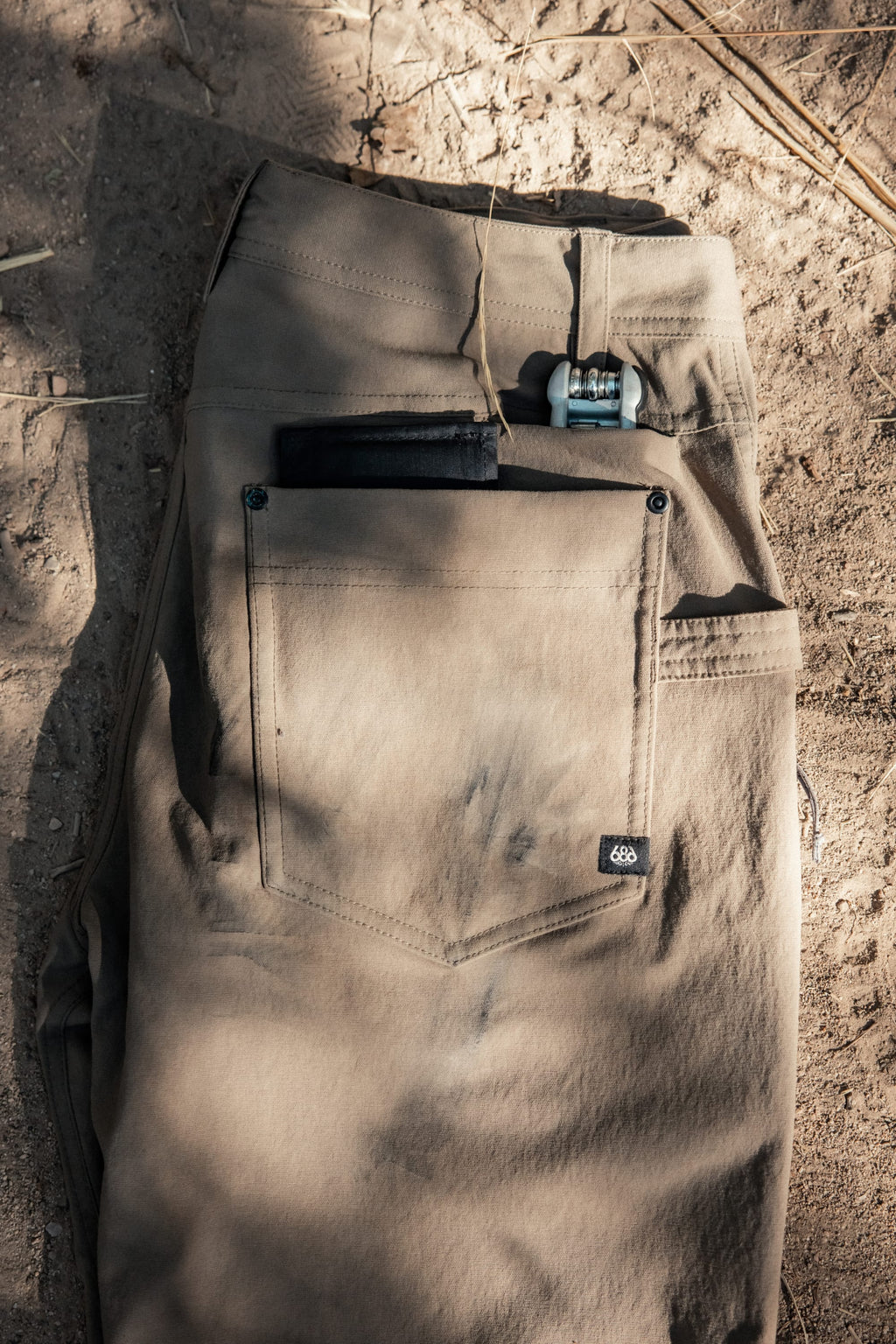 A pair of 686 Men's Unwork® Pant - Slim Fit in khaki lies folded on sandy ground with dry grass, black and blue items partially tucked into the back pocket.