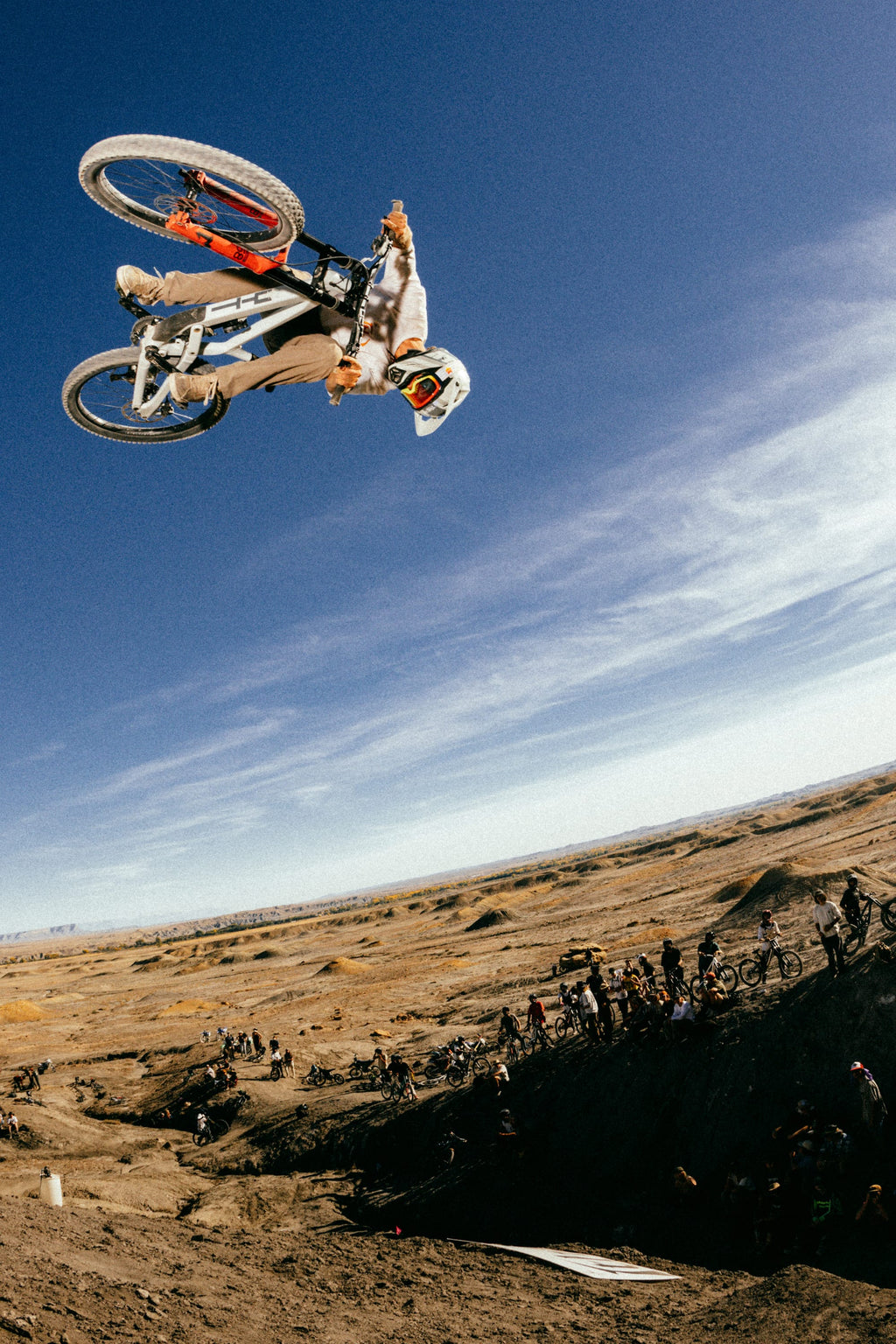 A motocross rider in 686 Men's Unwork® Pant - Slim Fit and a white helmet performs a high-flying trick above a dirt track as spectators watch from below under a clear blue sky.