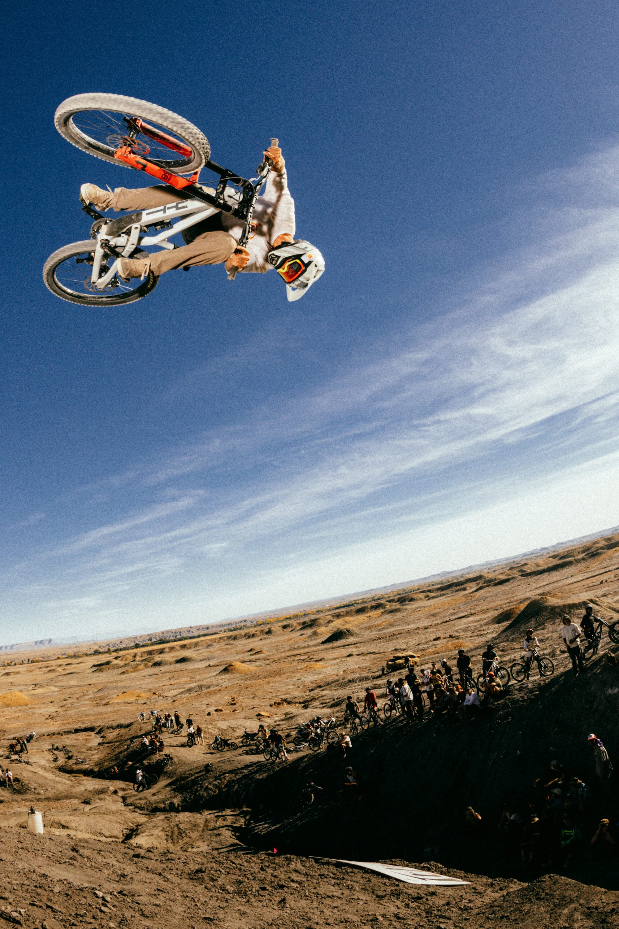 A motocross rider in 686 Men's Unwork® Pant - Slim Fit and a white helmet performs a high-flying trick above a dirt track as spectators watch from below under a clear blue sky.