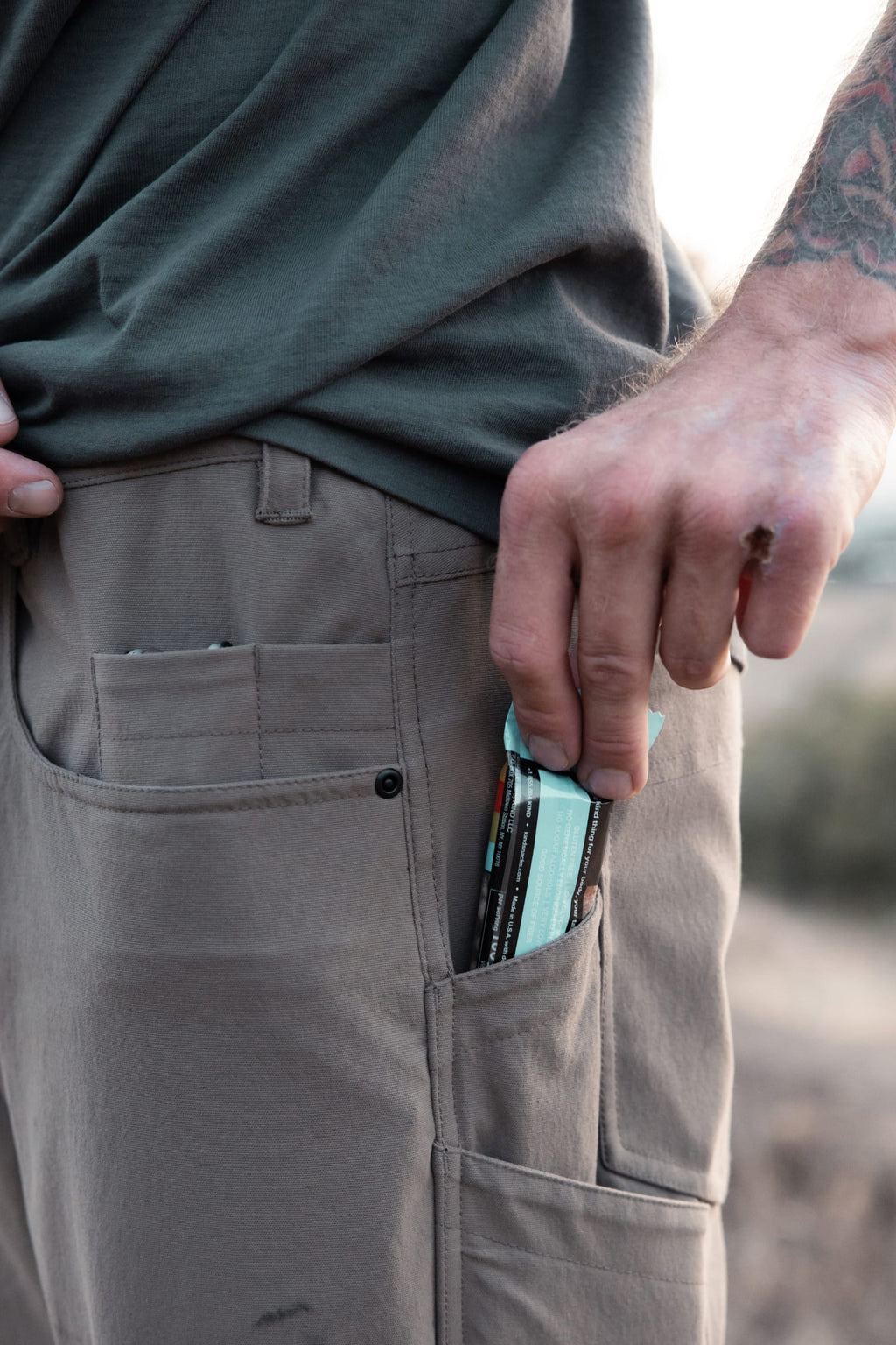 Wearing 686 Men's Unwork® Pant - Slim Fit and a green shirt, a person with a forearm tattoo puts two protein bars into the pants’ side pocket. Only their torso and hand are visible.