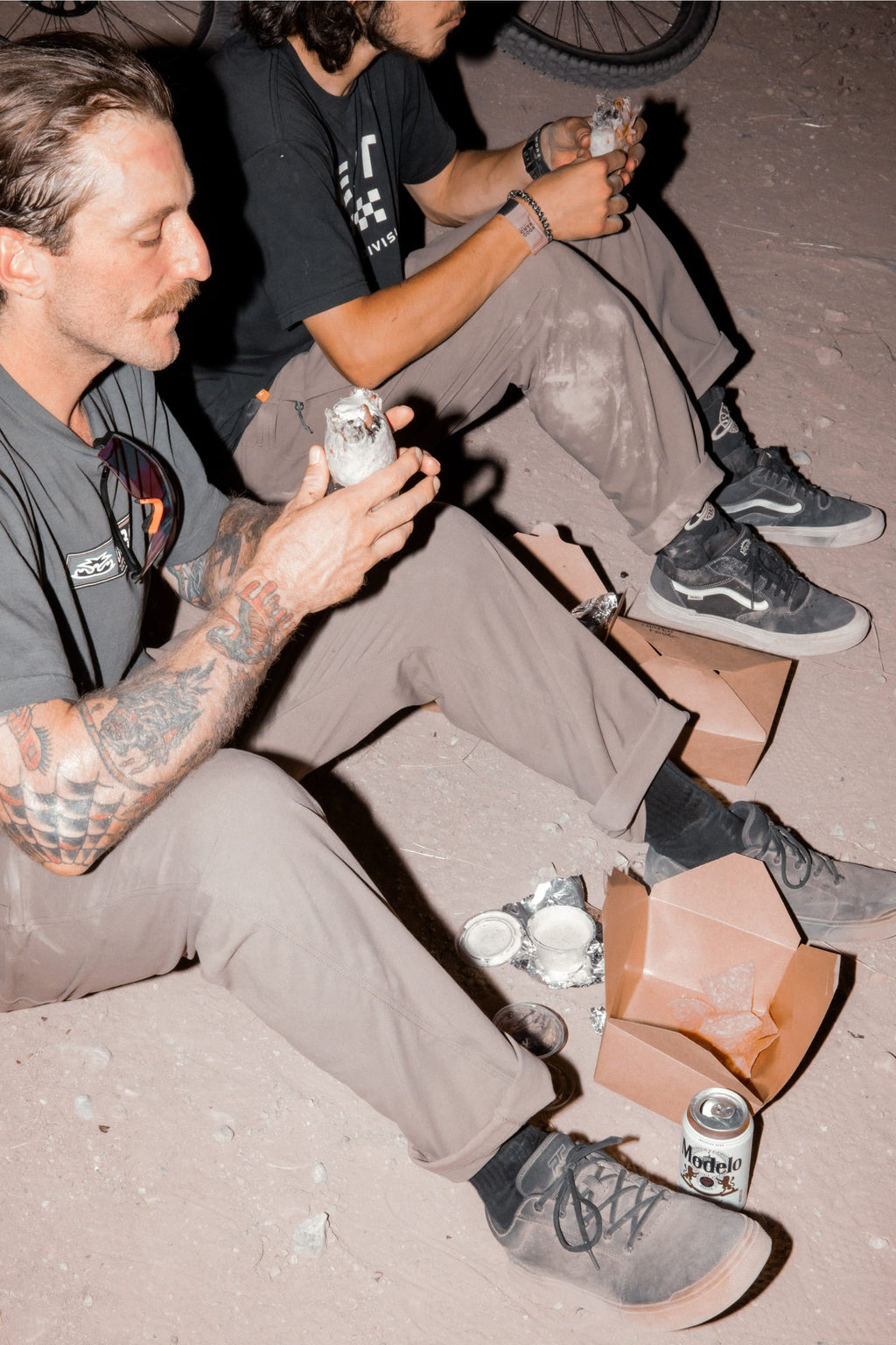 Two people sit on the ground at night, eating foil-wrapped burritos with takeout boxes and a can of Modelo nearby. Both wear casual clothing, sneakers, and 686 Men's Unwork® Pant - Slim Fit by 686—durable work pants perfect for the evening.