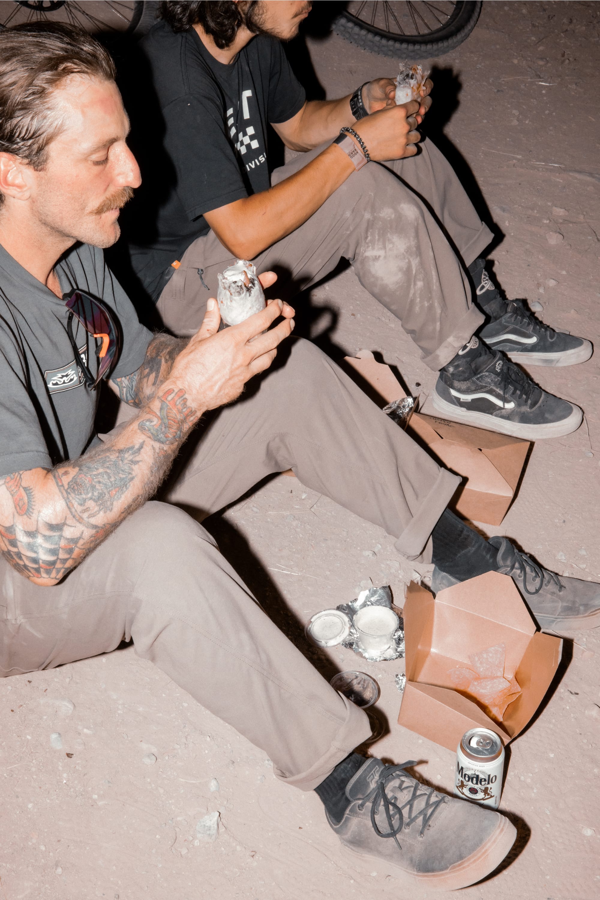 Two people sit on the ground at night, eating foil-wrapped burritos with takeout boxes and a can of Modelo nearby. Both wear casual clothing, sneakers, and 686 Men's Unwork® Pant - Slim Fit by 686—durable work pants perfect for the evening.