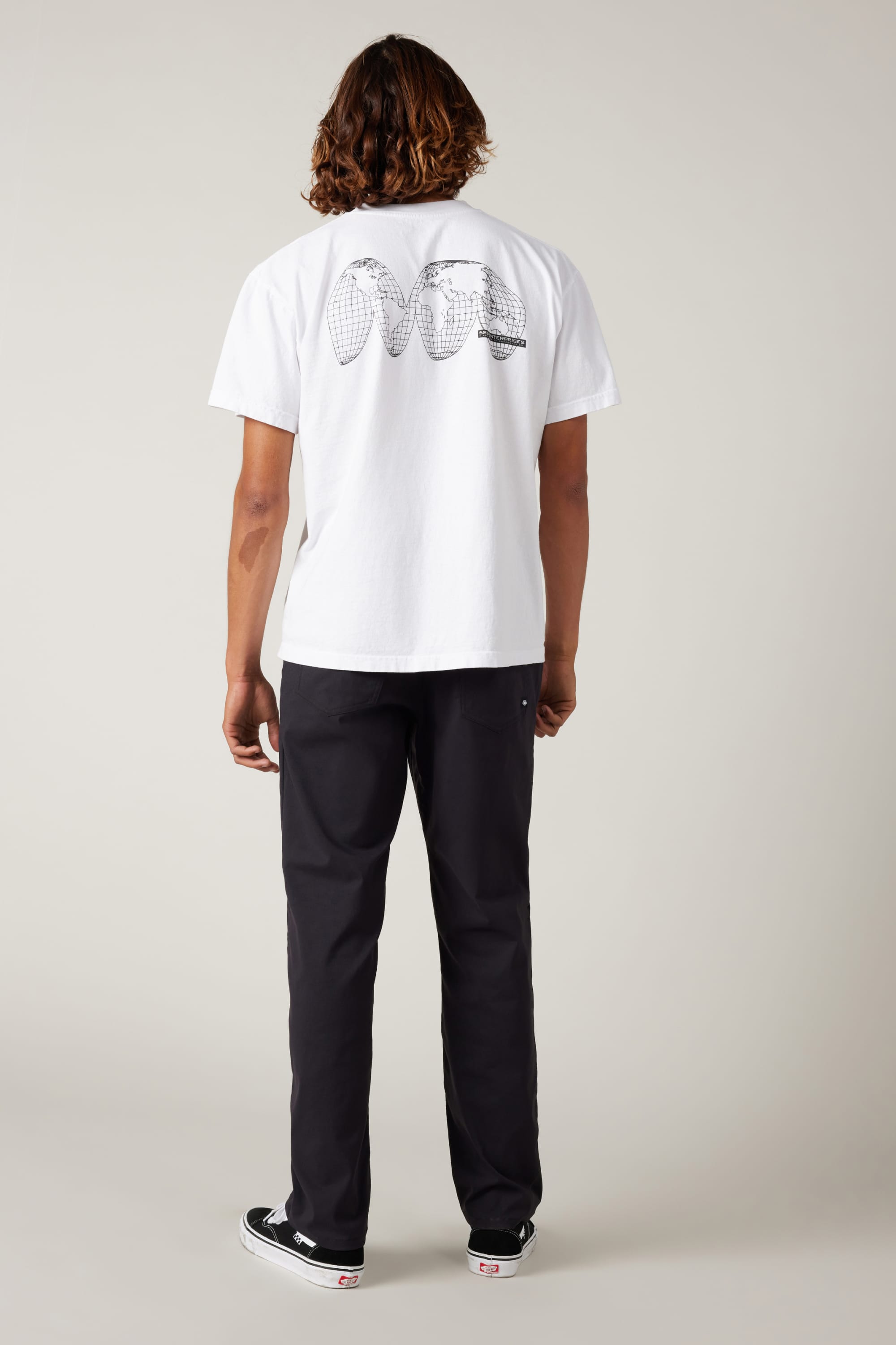 A person with shoulder-length hair stands facing away, wearing a white T-shirt and 686 Men's Everywhere® Pant - Straight Fit, highlighting their athletic legs. They also wear black slip-on sneakers with white soles against a neutral background.