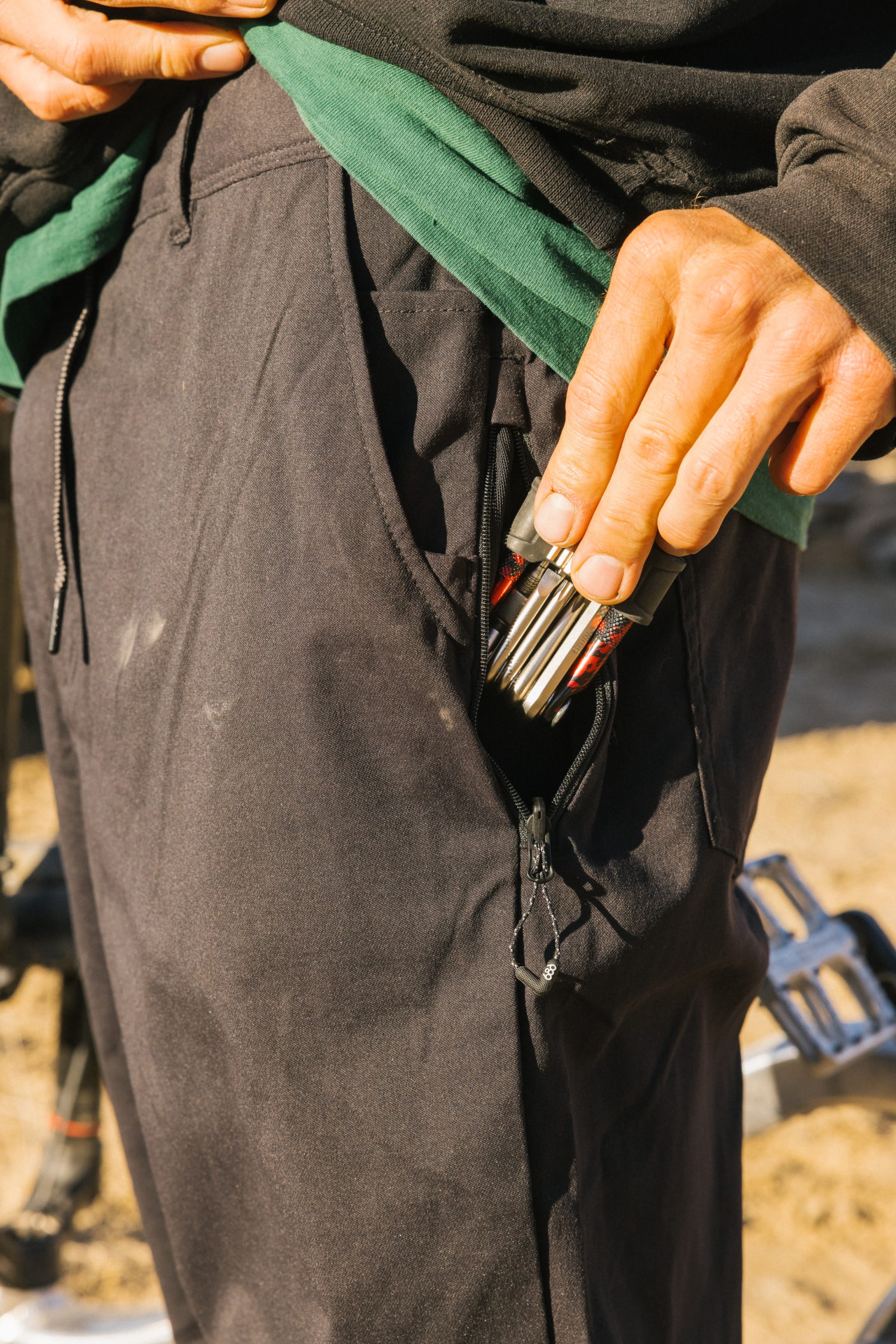 A hand slips a multitool into the zippered pocket of the 686 Men's Everywhere® Pant - Straight Fit. Set against an outdoor backdrop, these pants offer adventure-ready style, athletic fit, and on-the-go stain-resistant durability.