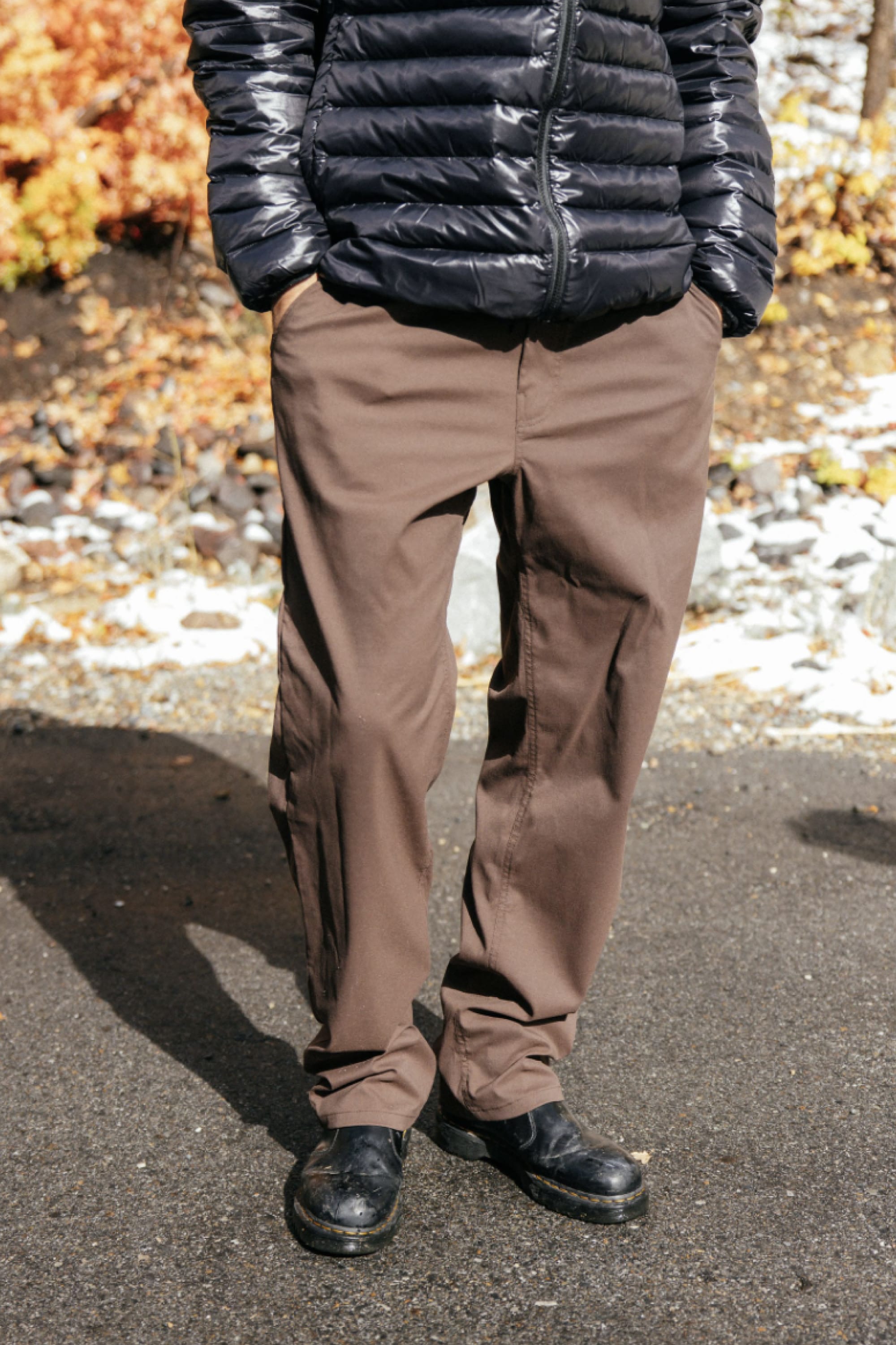 Outdoors on a paved surface, a person wears the 686 Men's Everywhere® Pant - Straight Fit, known for its stain resistance and athletic fit, paired with black shoes and a shiny black puffer. Their hands rest in their pockets, autumn foliage blurred in the background.