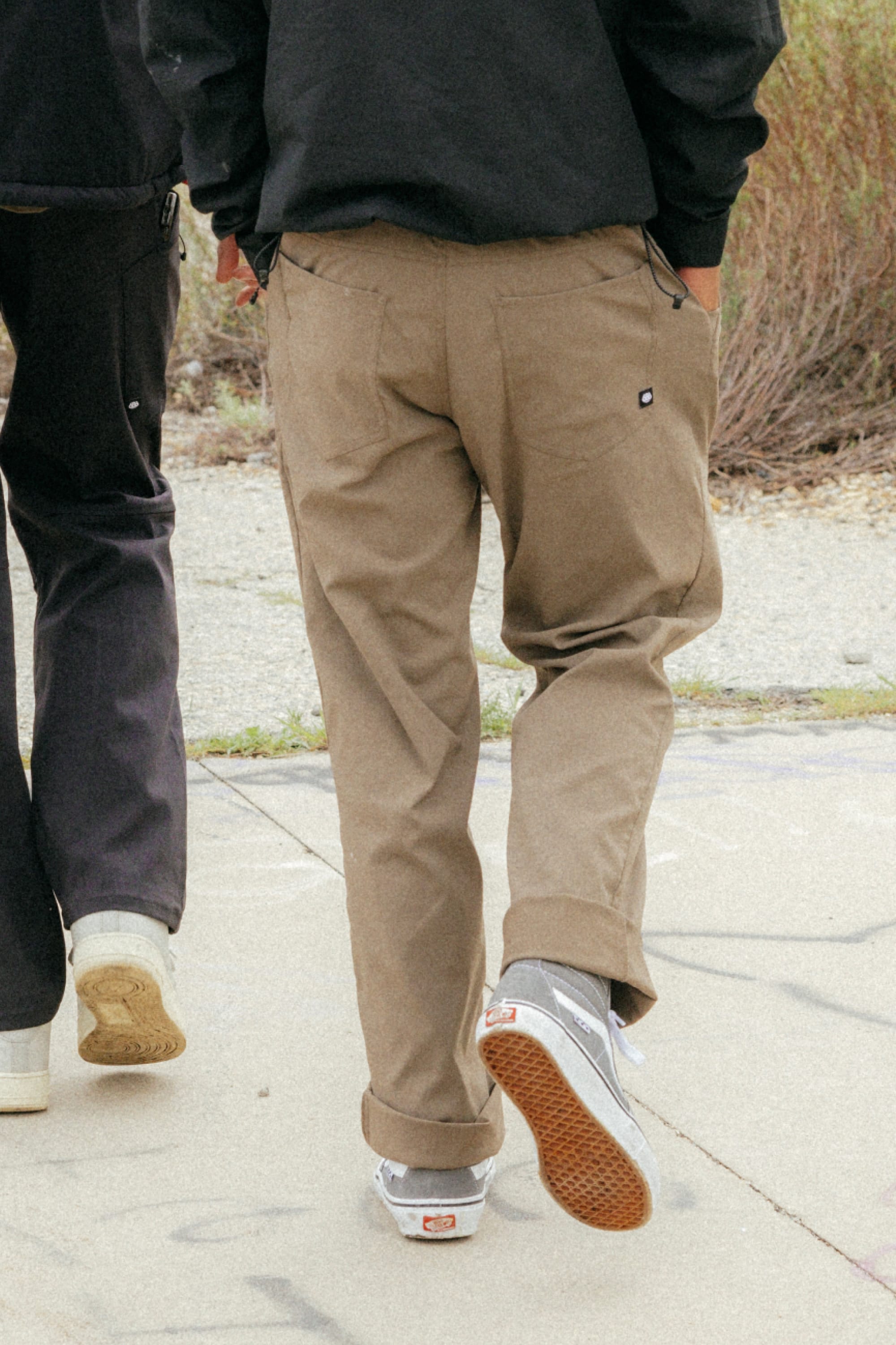 Two people walk outdoors on a paved path, showing just their legs—one in brown 686 Men's Everywhere® Pant - Straight Fit and gray sneakers, the other in dark pants with white sneakers. Grass and bushes fill the background.