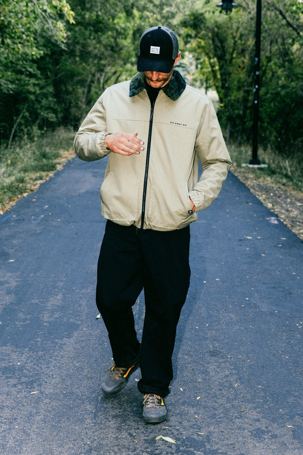 A person wearing a beige jacket, black 686 Men's Everywhere® Pant - Wide Fit, and a black cap stands on a paved path among trees, looking down with one hand in front of their chest and the other in their pocket.