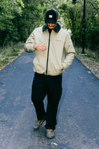 A person wearing a beige jacket, black 686 Men's Everywhere® Pant - Wide Fit, and a black cap stands on a paved path among trees, looking down with one hand in front of their chest and the other in their pocket.