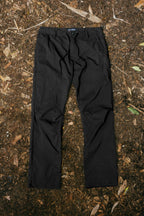 A pair of 686 Men's Everywhere® Pant - Wide Fit in black is laid flat on a forest floor strewn with dirt, dried leaves, and twigs.