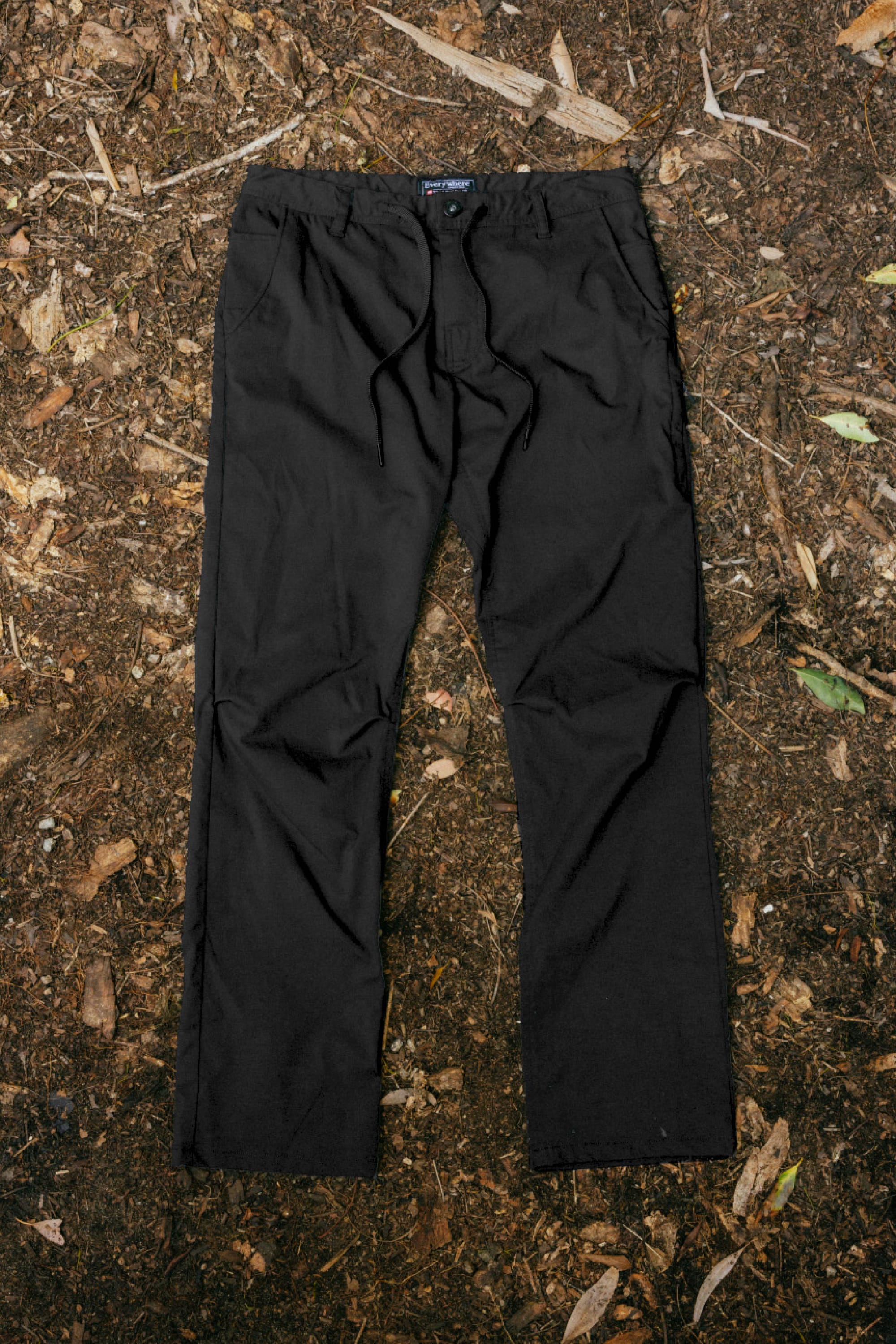 A pair of 686 Men's Everywhere® Pant - Wide Fit in black is laid flat on a forest floor strewn with dirt, dried leaves, and twigs.