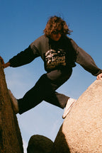 A person with shoulder-length hair, wearing a black sweatshirt and 686 Men's Everywhere® Pant - Wide Fit, balances with arms outstretched between two large rocks under a clear blue sky.