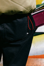 Close-up of a person wearing 686 Men's Everywhere® Pant - Wide Fit by 686, featuring a black-and-white logo tag and red cord, styled with wide legs, standing before a vibrant graffiti-covered wall.