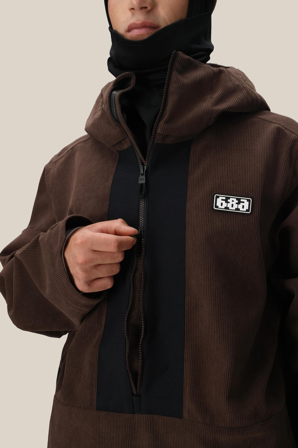 A person in the 686 Men's Ghost™ 2.5L Anorak—a dark brown, hooded jacket with black panels and a chest “6&8A” patch—partially unzips the rugged nylon shell, revealing a black inner layer while their face remains partially visible.