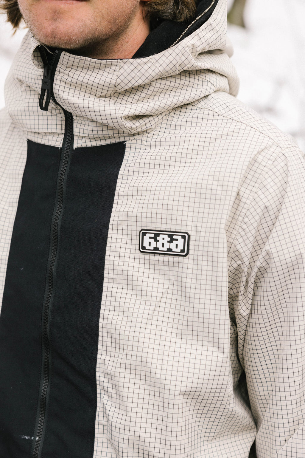 A person wears the 686 Men's Ghost™ 2.5L Anorak—a lightweight, shorter-fit shell with a light grid pattern, black vertical stripe, and 686 logo on the chest. Only their upper body and lower face are visible.