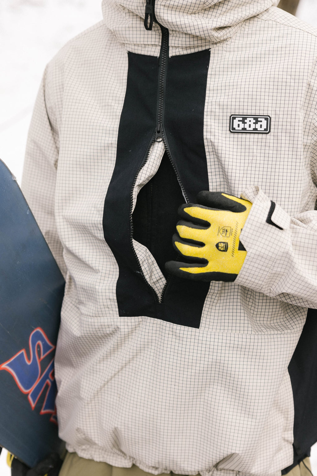 A person wearing the 686 Men's Ghost™ 2.5L Anorak holds the jacket with a yellow and black work glove, along with a blue snowboard featuring red lettering.
