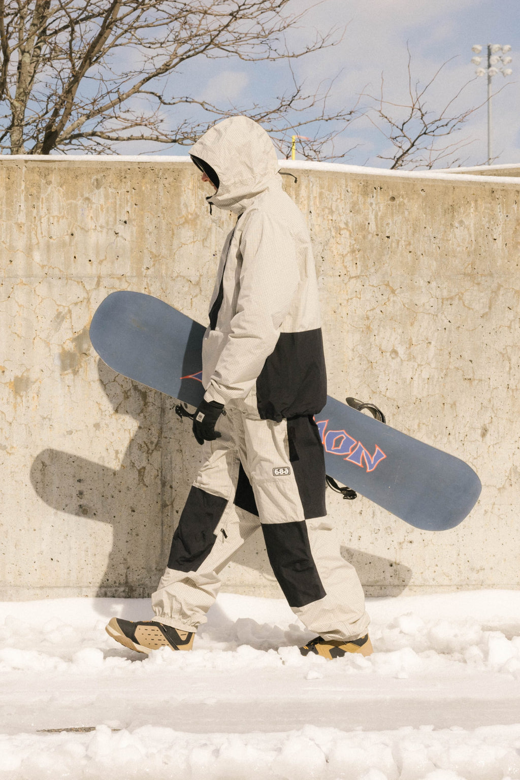 Wearing the 686 Men's Ghost™ 2.5L Anorak, a person in a beige and black snowsuit carries a blue snowboard past a concrete wall with bare branches and stadium lights behind, showing off the anorak’s lightweight shell and short fit.
